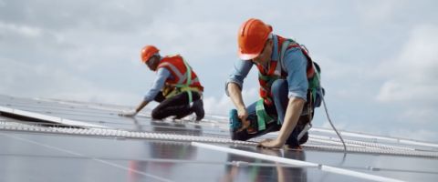 Two people in hard hats and work gear kneeling on solar panels.