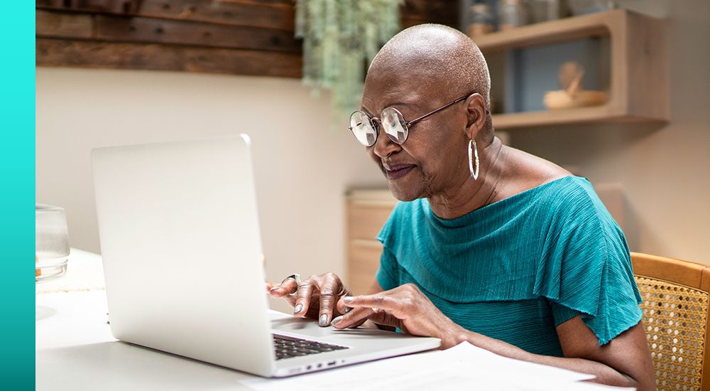 Woman in a blue shirt and glasses sitting at a table while working on a laptop computer