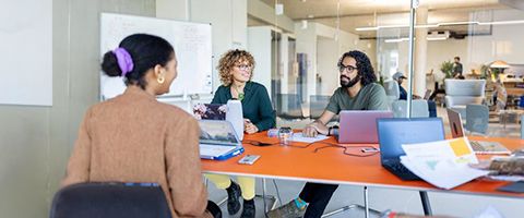 Three people sitting at an orange table in a conference room while working on laptops