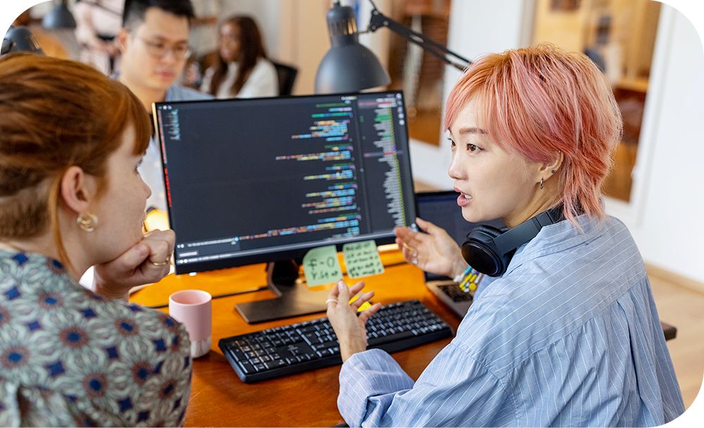 Person with pink hair and headphones sitting at a computer while having a conversation with the person next to them