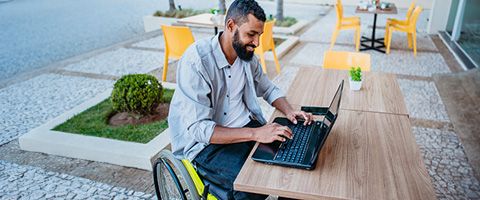 A man who uses a wheelchair works on a laptop outside in a café area 