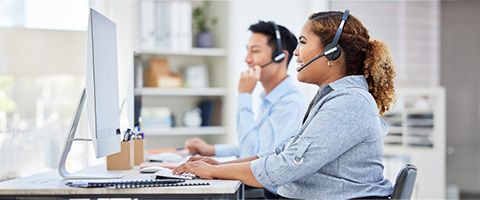 Two customer service representatives wearing headsets work at a desk 