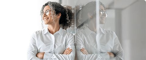 Businessperson poses  leaned against reflective glass with crossed arms and a smile.