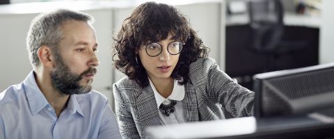 Two people sit together and review information on a desktop computer. 