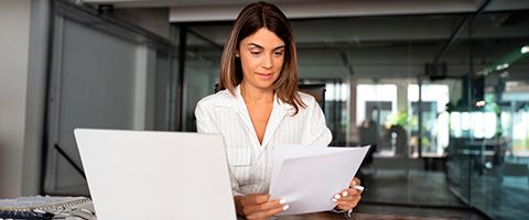 A businessperson sits at their desk in front of a laptop reviewing information in a packet in their hands