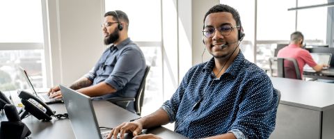 Two men in shirts sitting in a row of desks while wearing headsets and working on laptops