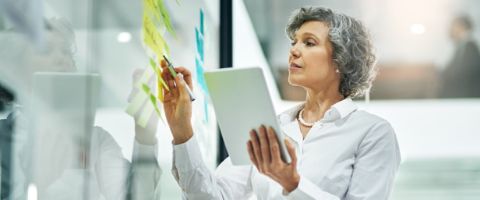 A person in a white button-up top holding a smart tablet and writing on a whiteboard. 