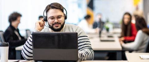 Man in a striped shirt sits and works on a laptop while wearing a headset and others work in the background