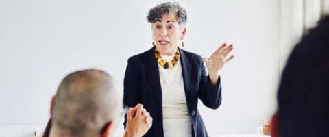 Woman in a dark coloured jacket standing while addressing people sitting at a table in front of her