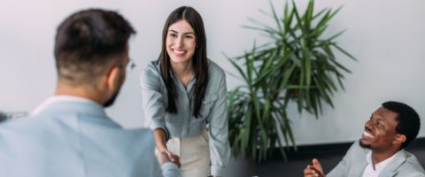 Woman in a grey shirt smiling while shaking hands with a person standing across from her while another man sits at the table