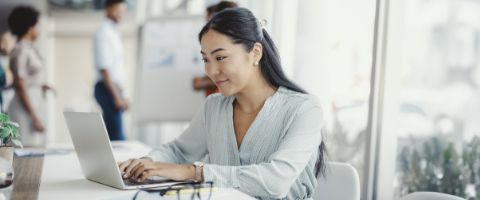 A woman focused on her laptop, seated at a desk in a modern office environment, surrounded by office supplies