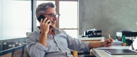 Man in a grey shirt and glasses sitting at a desk while talking on the phone