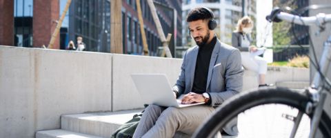 Person in business casual attire sitting on steps outside while wearing headphones and working on a laptop.