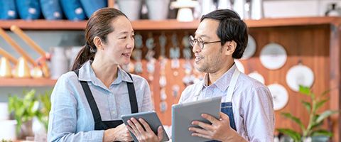 Two people wear aprons over collared shirts and talk while holding tablets.