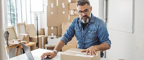 A person stands in front of a collection of shipping boxes and wears a collared button down, reading a shipping label on a box.