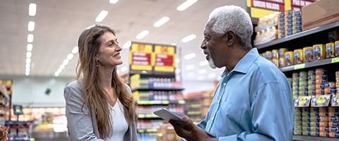 Two people stand in a grocery store and discuss something referencing information on a tablet. 