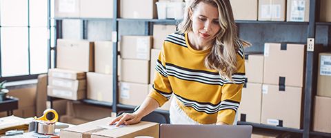 A person stands in a room full of shipping boxes and does something with a shipping label and laptop.