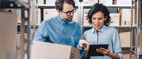 Two people stand in a room with shipping boxes and reference something between a shipping label and information on a tablet. 