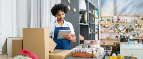 Person wearing a blue apron and holding a tablet next to boxes and merchandise in a retail setting.