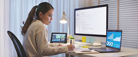 Person sitting a desk with two laptops open on either side, a large monitor in the middle and multiple papers for notes. 