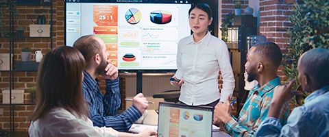 Businessperson standing in front of very large monitor discussion pie chart and other data to three colleagues. 