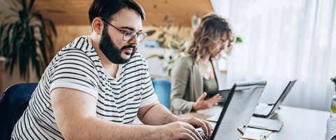 Man in a striped shirt and glasses sitting while working on a laptop in front of him with another person working next to him