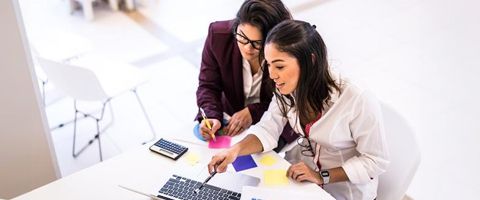 Two women work at a desk and discuss something on a laptop screen