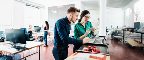 Two workers in casual business attire look at a laptop and talk