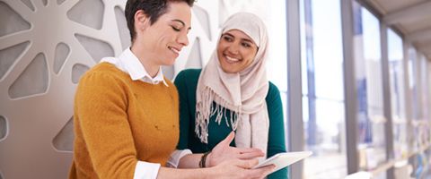 Two women smile and talk while one uses a tablet device