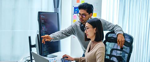 Person sitting at a desk typing responses as another manager points out next steps on the monitor. 