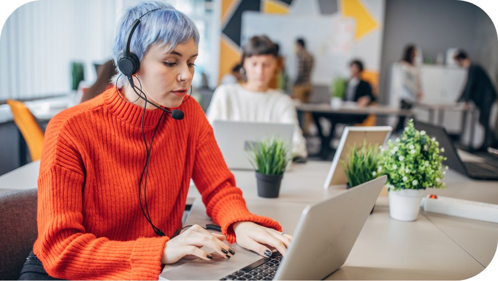 A person works on their laptop in an office setting, speaking into their headset.
