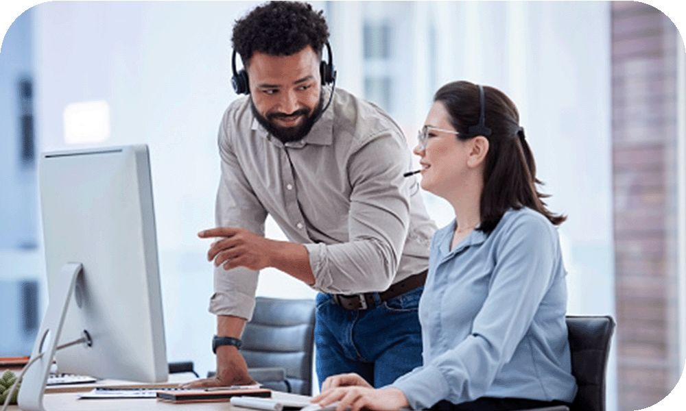 Man in a grey shirt and headset leaning over a desk talking with the woman in a blue shirt while working on her desktop computer