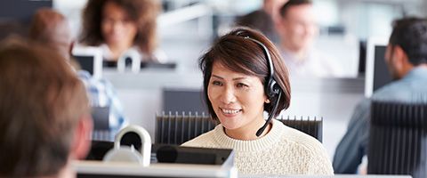 Woman in a white sweater sitting at a row of desks in front of a computer monitor while talking on a headset