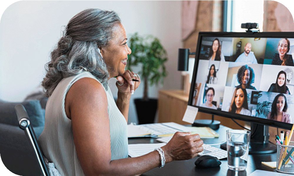 Woman in a white sweater sitting at a desk while looking at her desktop monitor open to a video call with nine people