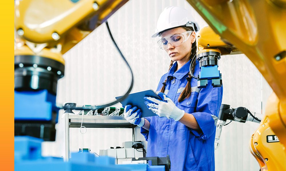 Person wearing a blue industrial uniform, white safety helmet and gloves, holds a tablet while standing between large yellow robotic arms in a factory or manufacturing setting.