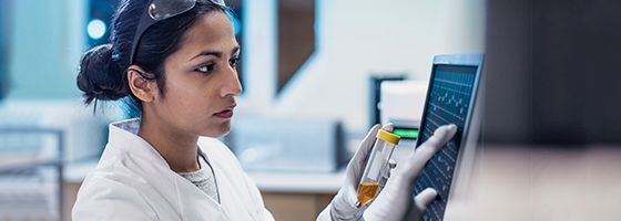 Frau in einem weißen Laborkittel, die sich an einer verschreibungspflichtigen Flasche festhält, während sie an einem Desktop-Monitor arbeitet