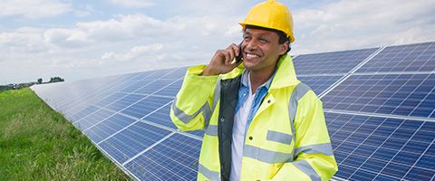 Worker in a hard hat and yellow safety vest smiles and talks on phone in front of a commercial solar panel