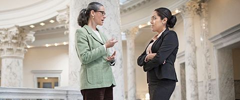 Two businesspersons in blazers talk with each other in a government building setting