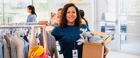 Worker in a blue shirt with a nametag and box of clothes in their hand stands near a clothes rack in a store