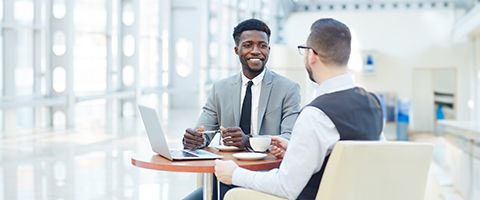 Two individuals seated at a round table in a bright office space, engaging in a discussion with an open laptop on the table