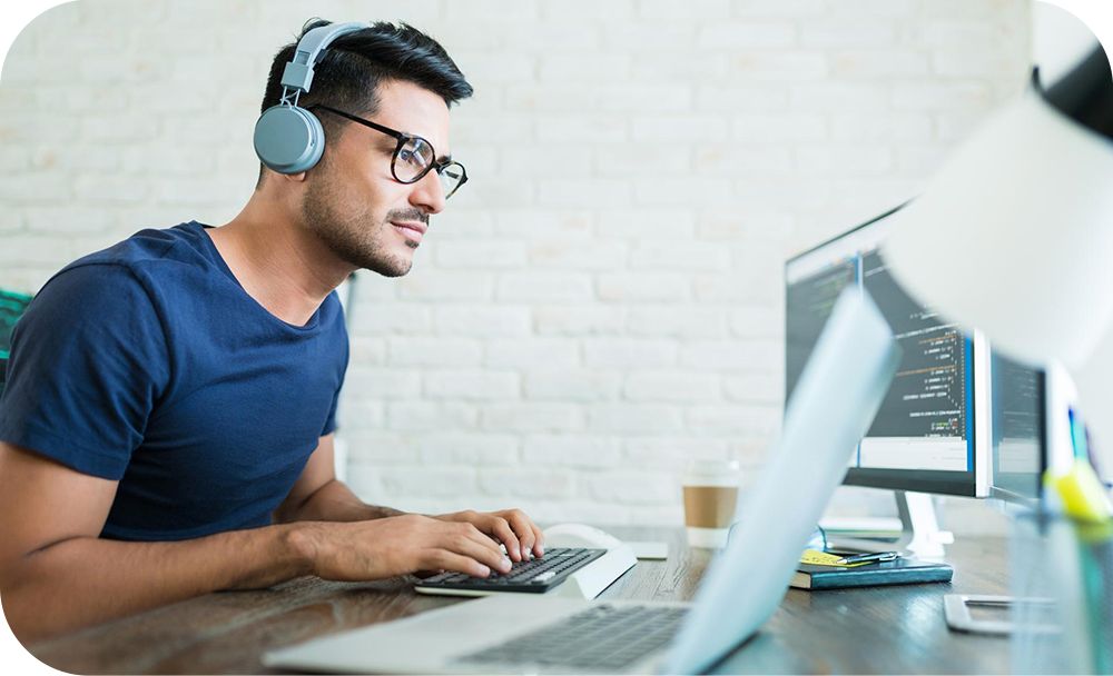 Person sitting at a desk and focused on a computer screen, wearing headphones with a coffee cup, notebooks and pens on the desk