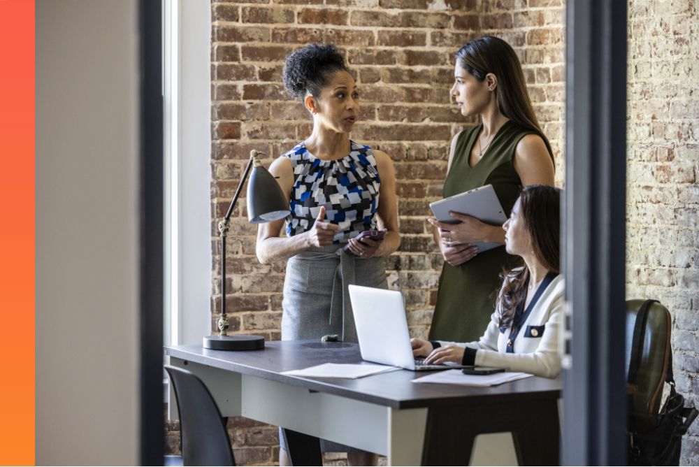 A person is seated at a desk with a laptop open in front of them while two other people are standing nearby engaged in a conversation 