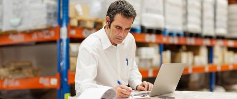 A person working on a laptop and writing on papers in a warehouse setting.