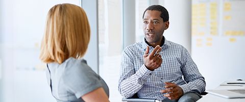 Two people in business casual attire talking and sitting at desks in an office setting.