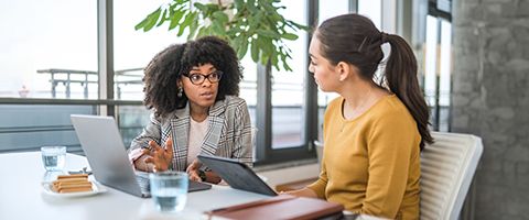 Two people in business casual attire talking over laptops at a desk in an office setting. 