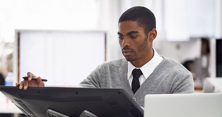 Geschäftsmann, der in einem hell erleuchteten Büroraum sitzt und einen Stift hält, während er an einem Computermonitor arbeitet