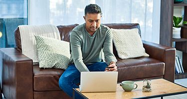 Man sitting and holding a credit card on a brown leather couch while buying something on his laptop