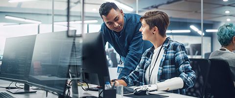 Two people seated at desktop computers in a modern workplace with a third person partially visible in the background