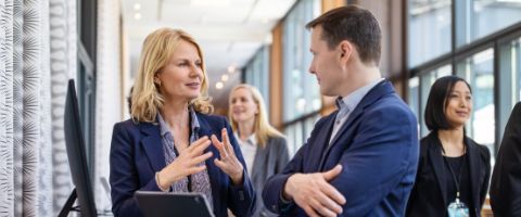 Two businesspeople standing and talking in a room with other people standing and chatting in the background