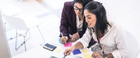 A person sits at a desk looking at documents while another leans over and writes something on a sticky note